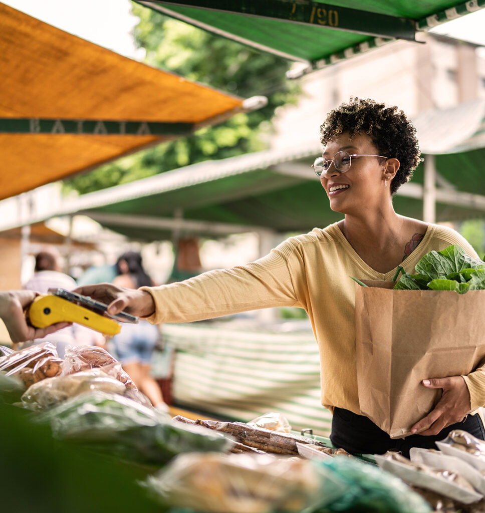 Eine Frau steht an einem Marktstand, der sich draußen befindet. In einem Arm hält sie eine Tüte. Der andere Arm ist ausgestreckt, mit diesem hält sie ihr Handy auf ein Zahlungsgerät. Das Zahlungsgerät wird von einer zweiten Person gehalten.