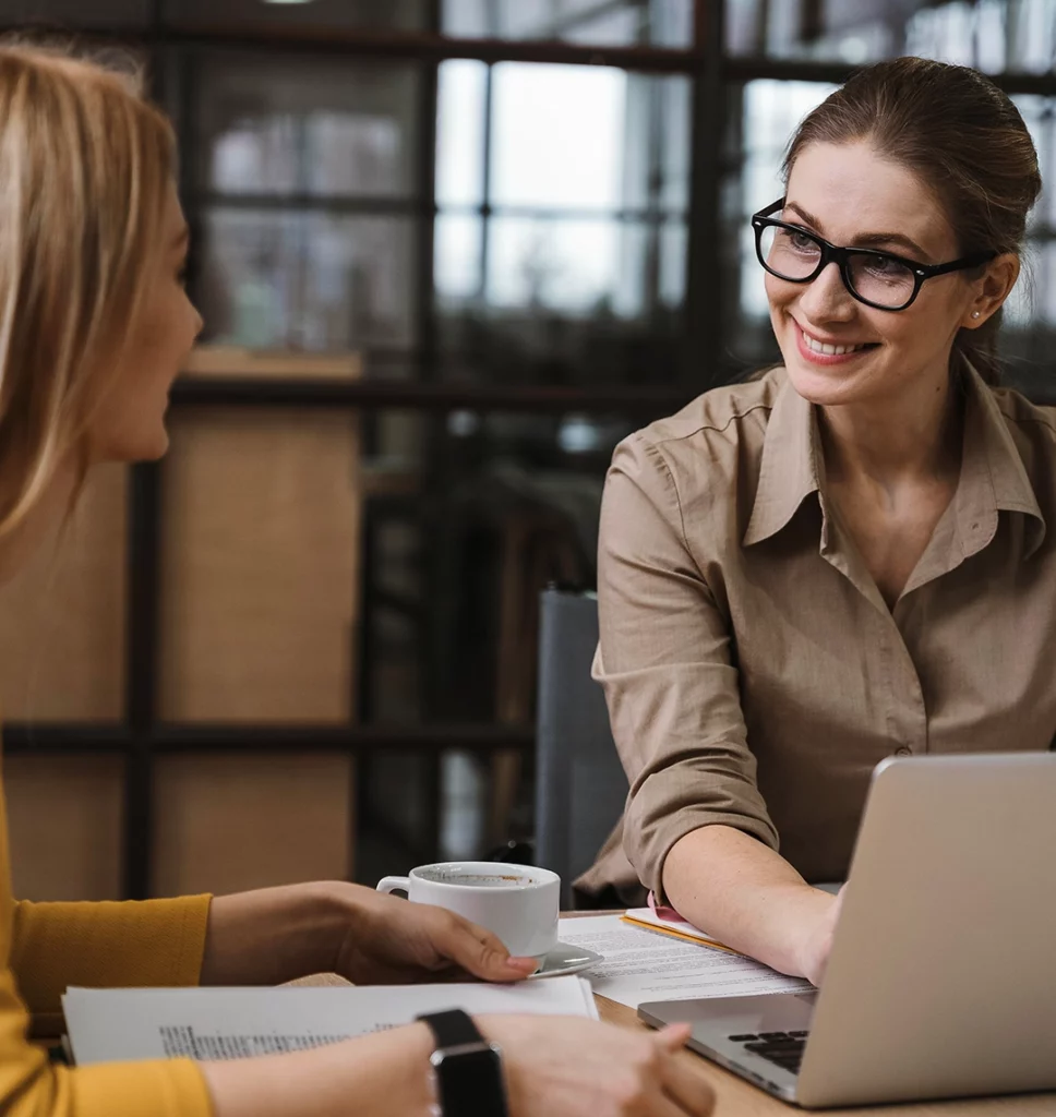 Zwei junge Frauen sitzen an einem Tisch und lächeln sich an. Neben ihnen steht ein Kaffee und ein Laptop.
