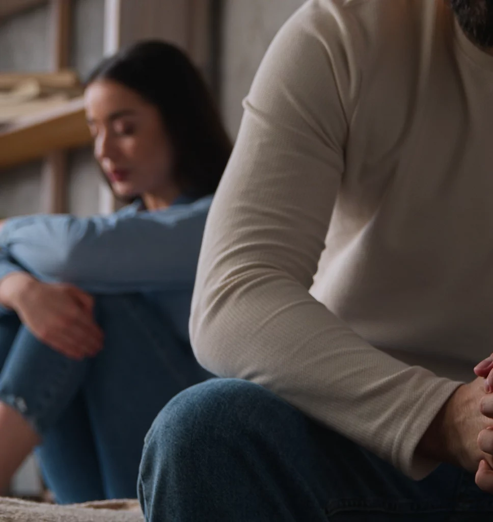 Eine Frau sitzt hinter einem Mann. Sie ist verschwommen. Sie umarmt ihre eigenen Beine und schaut traurig und nachdenklich. Von dem Mann sieht man nur ein Bein und seine Schulter. Die Stimmung des Bildes wirkt angespannt.