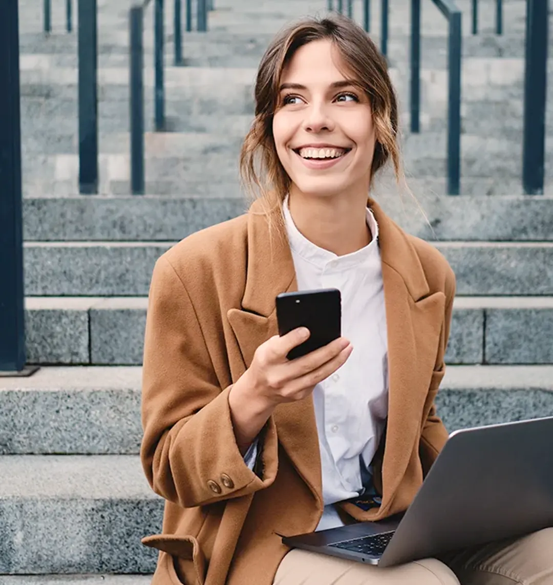 Eine Frau sitzt auf einer Treppe. Sie hält ihr Smartphone in der Hand und hat einen Laptop auf dem Schoss. Sie lächelt.