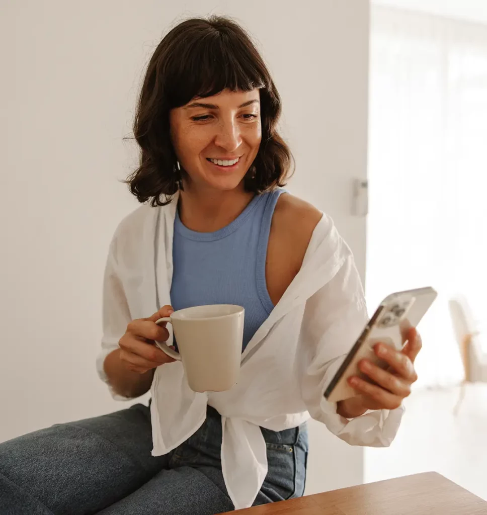 Eine Frau sitzt auf einem Stuhl in einem hellen Raum. Sie hält eine Tasse und ein Smartphone in der Hand und schaut auf das Smartphone. Die Atmosphäre ist entspannt, die Frau lächelt und sieht glücklich aus.