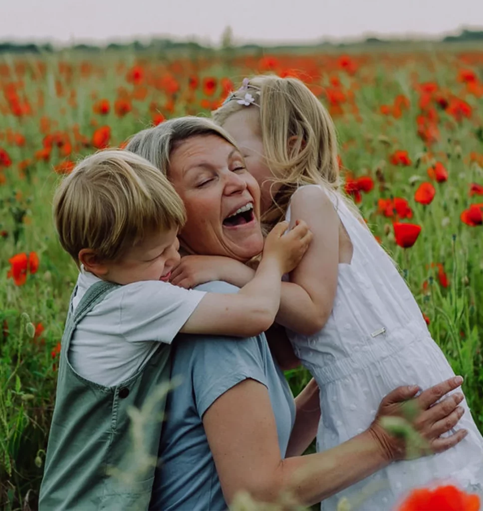Cathrin Clausnitzer sitzt mit ihren beiden Kindern vor einer Mohnwiese. Die Familie steht im Vordergrund, sie umarmen sich herzlich lachend.