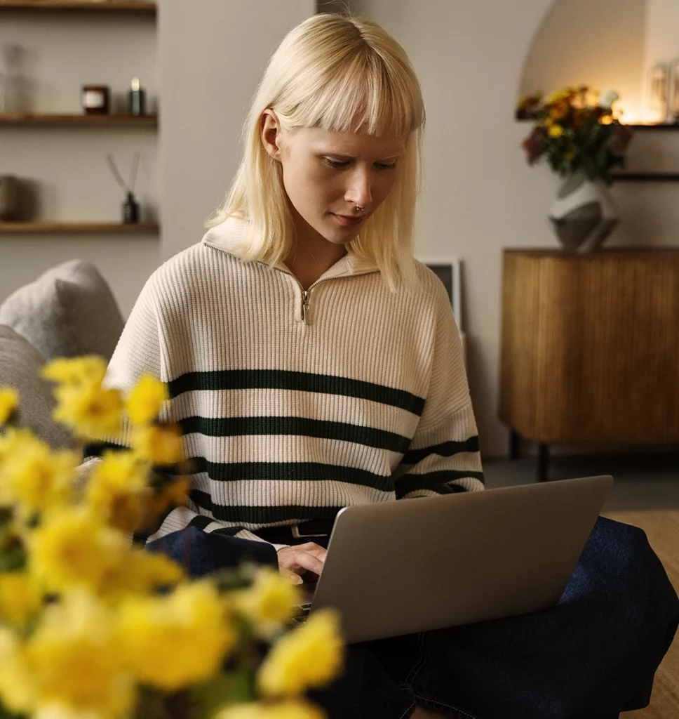 Eine junge Frau sitzt in ihrem Wohnzimmer am Laptop. Neben ihr stehen gelbe Blumen, die Atmosphäre ist ruhig.