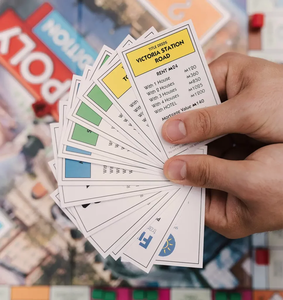 Im Hintergrund ist verschommen ein Monopoly-Spielbrett. Fokussiert wird auf Hände im Vordergrund, welche verscheidenen Straßenkarten aufgefächert halten