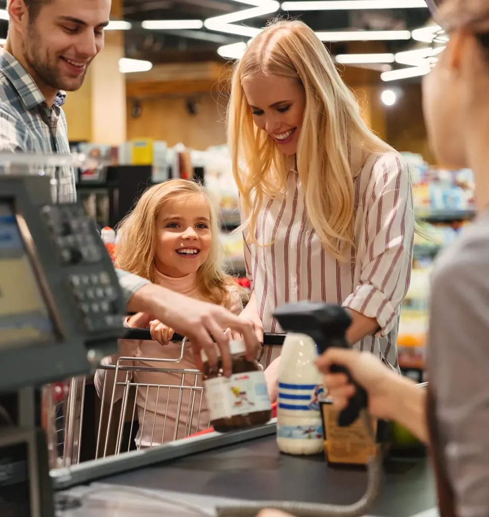 Junges Paar mit Tochter steht an einer Supermarktkasse und die Verkäuferin scannt die Produkte