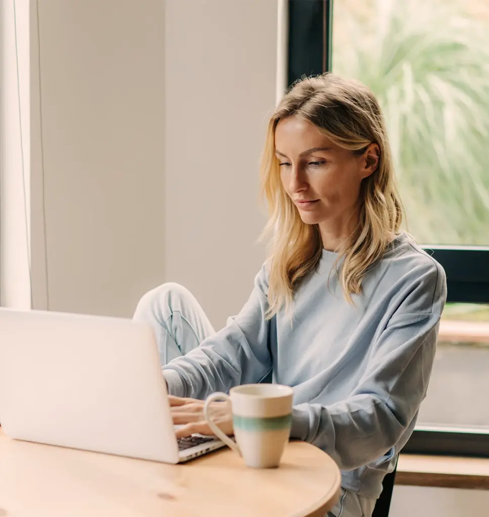 Eine Frau in einem leichten Pullover sitzt an einem Tisch und tippt auf einem Laptop. Daneben steht eine Tasse, in einem hellen Raum mit einem großen Fenster.